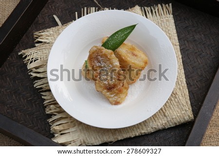 Fried bananas on a white ceramic plate and garnished with pandanus leaf. Underlined with folded straw mat and placed on dark color bamboo tray.