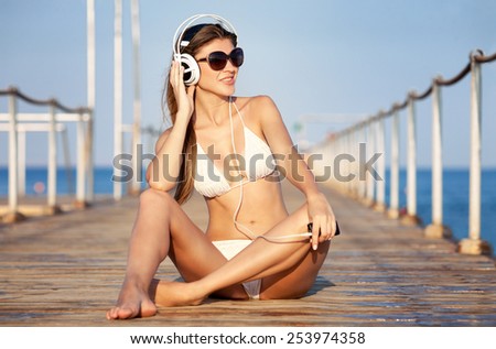young woman in headphones sitting on beach and enjoying beautiful sunset over the sea, rear view