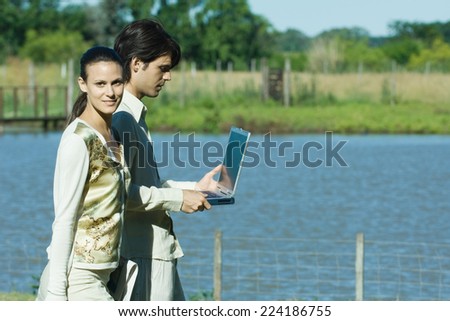 Man and woman standing by lake, man holding laptop, woman looking at camera