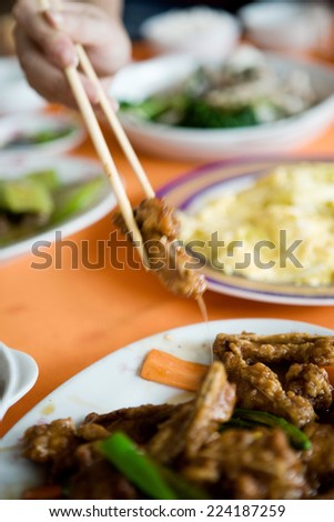 Several prepared dishes on table, person reaching for meat with chopsticks, cropped view