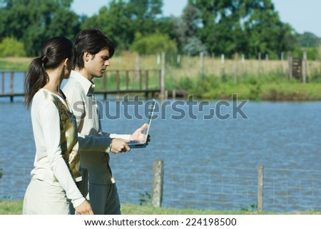 Man and woman standing by lake, man holding laptop