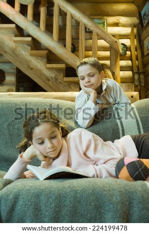Girl on couch reading book, boy looking over shoulder