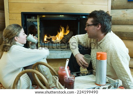 Young man and teenage girl sitting by fireplace, drinking hot beverages, looking at each other