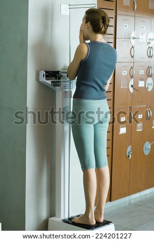 Young woman standing on scale in locker room, covering mouth