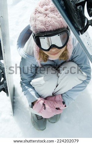 Preteen girl sitting on snow underneath propped up snowboards, high angle view
