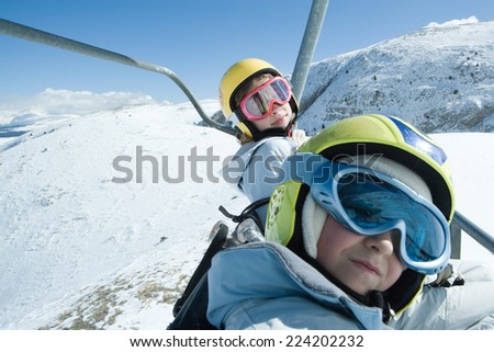 Two young friends on ski lift, both wearing ski goggles, one looking at camera