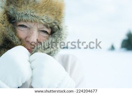Senior woman pulling flaps of fur hat over face, in snowy landscape, close-up