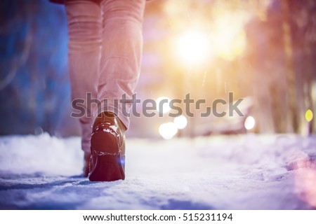 Legs of woman walking in winter park evening. Girl boots walking snow weather. Closeup of winter shoes. Blurred lens flare background with copy space area for a text.