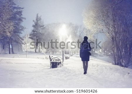 Snowfall in the city park. Woman walking down by alley. Night landscape with winter falling snowflakes. Winter snowfall scene. Severe weather in the winter park snowfall trees, benches.