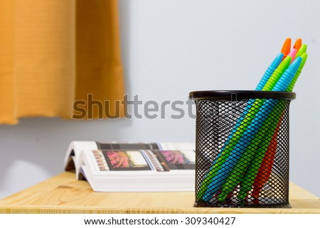 Colorful pen in metal pen pot with the book on the wood table