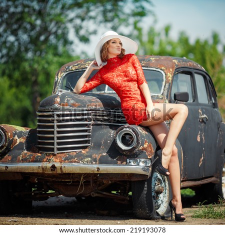 Beautiful young woman looks sexy, comes against the backdrop of an old black car in a red dress. Girl in red dress holding a white hat. Image of a woman who looks away.