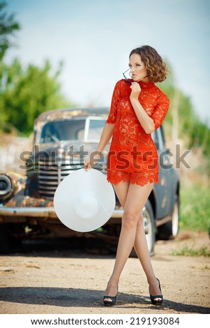 Beautiful young woman looks sexy, comes against the backdrop of an old black car in a red dress. Girl in red dress holding a white hat. Image of a woman who looks away.
