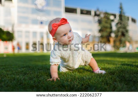 Little baby playing on the grass in a city park. Joyful baby with red bow crawls on all fours on the grass. Children\'s yoga. The kid looks overlooking pine.