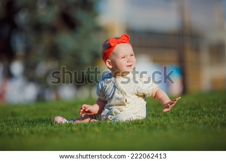 Little baby playing on the grass in a city park. Joyful baby with red bow crawls on all fours on the grass. The kid looks overlooking pine.