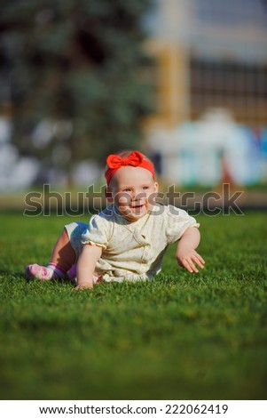 Little baby playing on the grass in a city park. Joyful baby with red bow crawls on all fours on the grass. The kid looks overlooking pine.