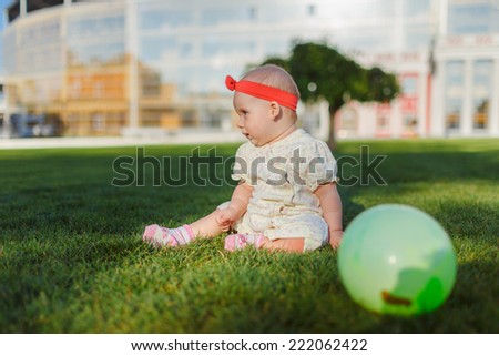 A young child plays with a ball on the grass in a city park. Joyful baby with red bow crawls on all fours on the grass. The kid looks overlooking pine.