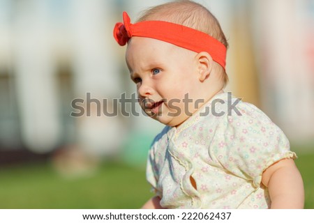 Little baby playing on the grass in a city park. Joyful baby with red bow crawls on all fours on the grass. The kid looks overlooking pine. Close portrait of a baby