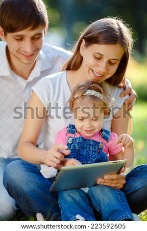 Happy family of three people relaxing in a city park. Father shows in tablet funny pictures. Family sitting on grass and looking at the tablet. Happy family concept of the good life.