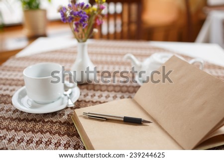 Pen and notebook on the table in a cafe, standing next to a cup of tea, teapot and vase with a bouquet of flowers