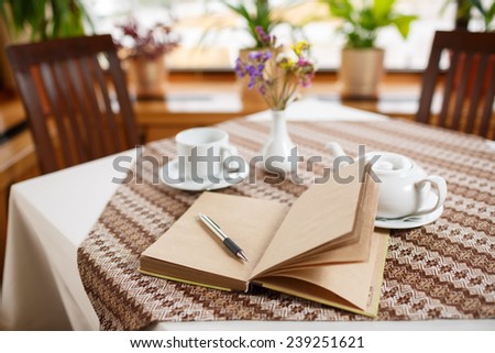 Ballpoint pen and notebook on the table in a cafe, standing next to a cup of tea, teapot and vase with a bouquet of flowers