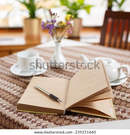 Pen and notebook on the table in a cafe, standing next to a cup of tea, teapot and vase with a bouquet of flowers, top view