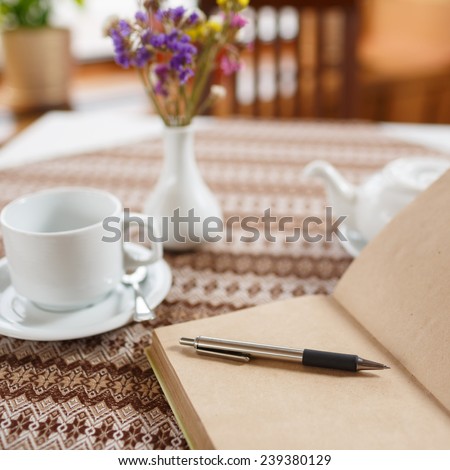 Ballpoint pen and notebook on the table in a cafe, standing next to a cup of tea, teapot and vase with a bouquet of flowers