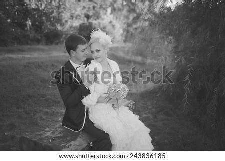Black and white photo. Beautiful young couple bride and groom, the European appearance, sitting on a bench on a sunny day outdoors. The groom embraces the bride\'s shoulders while sitting on the bench