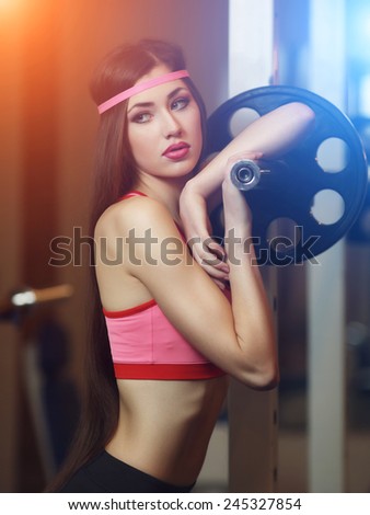Sexy brunette with very long hair and sporting appearance posing in the gym under the rays of blue and orange light. Young fitness woman in pink t-shirt trainers at the gym.