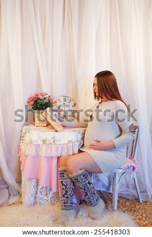 Pregnant woman reading a book while sitting at a table on which stands a vase with pink flowers and watches