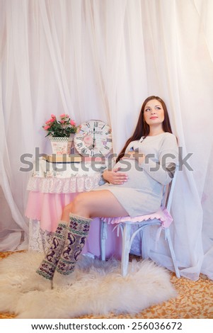 Beautiful pregnant woman sitting on a chair and hugs her belly. Young pregnant brunette, European, stroking her belly sitting on a chair next to the table is a vase of flowers and watches