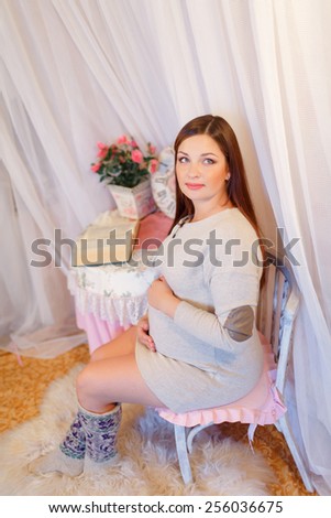 Beautiful pregnant woman sitting on a chair and hugs her belly. Young pregnant brunette, European, stroking her belly sitting on a chair next to the table is a vase of flowers and watches