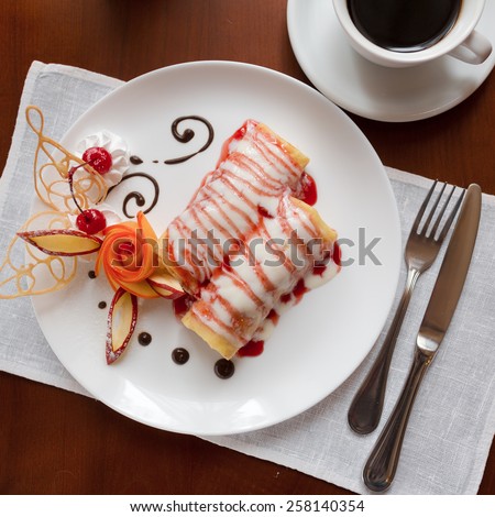 Pancakes with cherry jam beautifully decorated with flowers of fruit and chocolate on a white plate, which stands on a wooden table near the window in the interior cozy cafe. Shallow depth of field.
