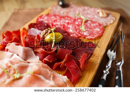 Variety of meats, sausages, salami, ham, olives, laid out on a wooden board close-up, horizontal, are next to a knife and fork