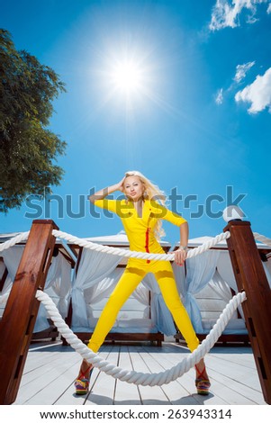 Sexy blonde with long straight hair and plump lips posing in a yellow suit in the sun, blue sky and white developing material