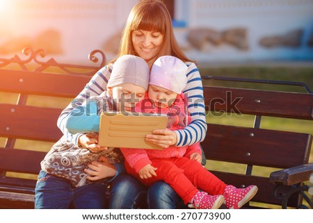Happy family of three, mother, son and daughter sitting on a bench in the afternoon in the city park and are holding a tablet, a beautiful spring weather, the sun is shining