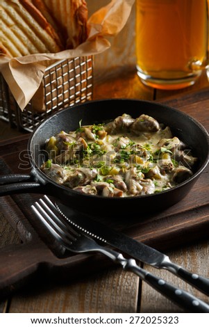 Roast with mushrooms, garlic sauce and chopped greens served in a frying pan on a wooden board, standing next to a glass of beer and golden pieces of bread