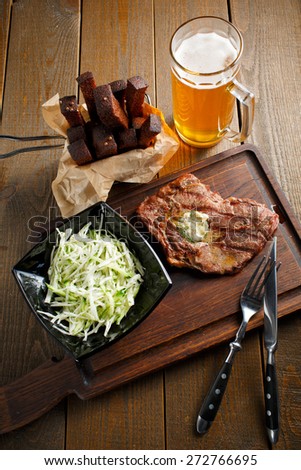 Delicious fried steak with butter and herbs, black plate of salad with fresh cabbage, beer and rye toast on dark wooden background