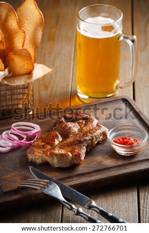 A delicious steak with chili sauce and chopped onion rings, chips of white bread in a metal basket and a glass of beer on a dark wooden background
