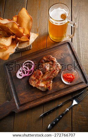 A delicious steak with chili sauce and chopped onion rings, chips of white bread in a metal basket and a glass of beer on a dark wooden background