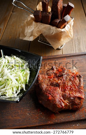 A delicious steak with red chili pepper, black plate of salad with fresh cabbage and rye toast croutons on wooden brown background