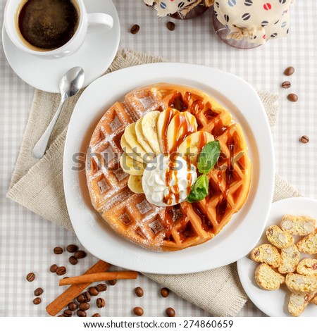 Belgian waffles with bananas and whipped cream, decorated with caramel sauce and powdered sugar on the table with a cup of coffee, coffee beans with cinnamon and two glass jars