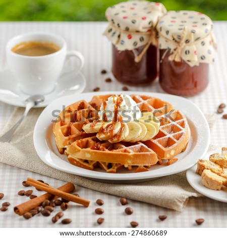 Belgian waffles with bananas and whipped cream, decorated with caramel sauce and powdered sugar on the table with a cup of coffee, coffee beans with cinnamon and two glass jars