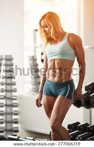 Young muscular woman bodybuilder with white hair and tanned body posing in white gym at the counter with dumbbells