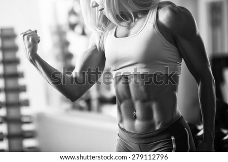 Black and white photo of a strong woman. Brutal blond with a muscular, tanned body, straining biceps and abdominal muscles against the window in the gym, part of the body, horizontally frame