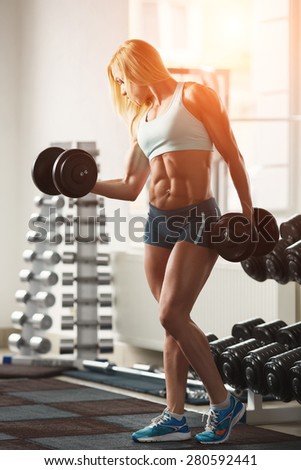 Strong woman bodybuilder with white hair and tanned body pumps up the muscles lifting dumbbells in the gym. Vertical frame with space for text
