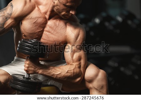 A very strong young guy bodybuilder, doing exercises with dumbbells in the gym. Fitness muscular body on dark background