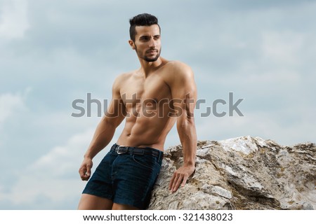 Muscular male torso on a background sky. Athletic and muscular man with naked torso stands on top of a mountain. Handsome young guy posing sporty appearance on the sky background, looking away