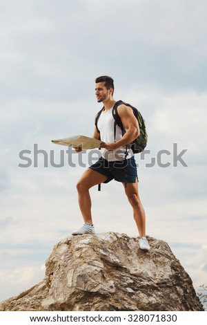 Hiker with a backpack standing on top of the mountain holding a map. Man with map exploring wilderness on trekking adventure. Tourist on the background of the sky