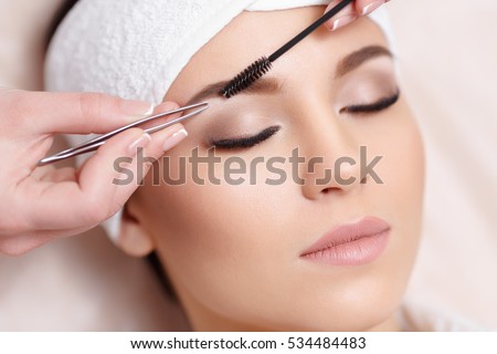 Permanent makeup. Beautiful young woman gets eyebrow correction procedure. Young woman tweezing her eyebrows in beauty saloon. Young woman plucking eyebrows with tweezers close up