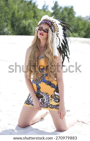 Young blonde woman in costume of American Indian sits on the sand
